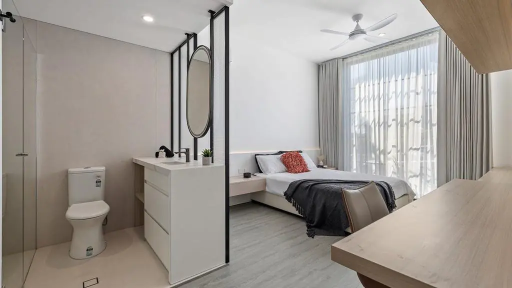 Modern bedroom with an ensuite bathroom featuring a toilet and sink, a bed with white and gray bedding, a ceiling fan, sheer curtains, and wooden furniture. Bright, minimalist decor with natural light.
