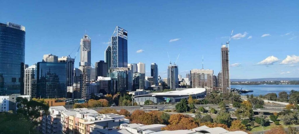 A Australian city skyline featuring modern skyscrapers, mid-rise buildings, and a large convention center, with a river and trees in the foreground under a clear blue sky.