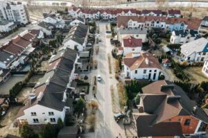 Aerial view of a suburban neighborhood with rows of houses and parked cars along a central street on a sunny day. Trees and small yards surround the homes, and a river is visible in the background.