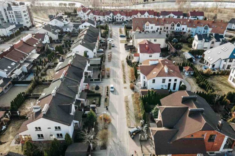 Aerial view of a suburban neighborhood with rows of houses and parked cars along a central street on a sunny day. Trees and small yards surround the homes, and a river is visible in the background.