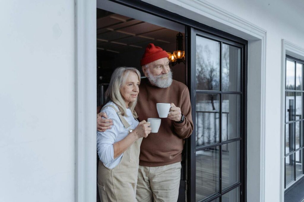 An older couple stands together on a porch, holding white mugs and looking outside. The woman wears a light blouse and beige apron, while the man wears a red beanie and brown sweater. They appear relaxed and content.