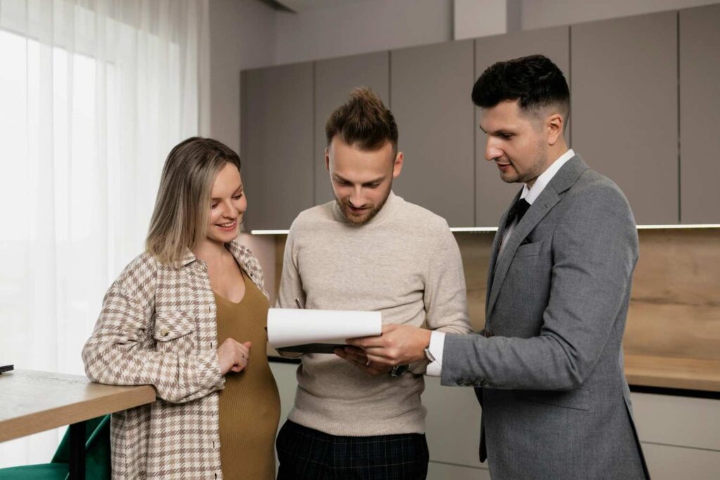 A man in a suit shows documents to a smiling couple in casual clothes, standing together in a modern kitchen with light-colored cabinets and a large window.