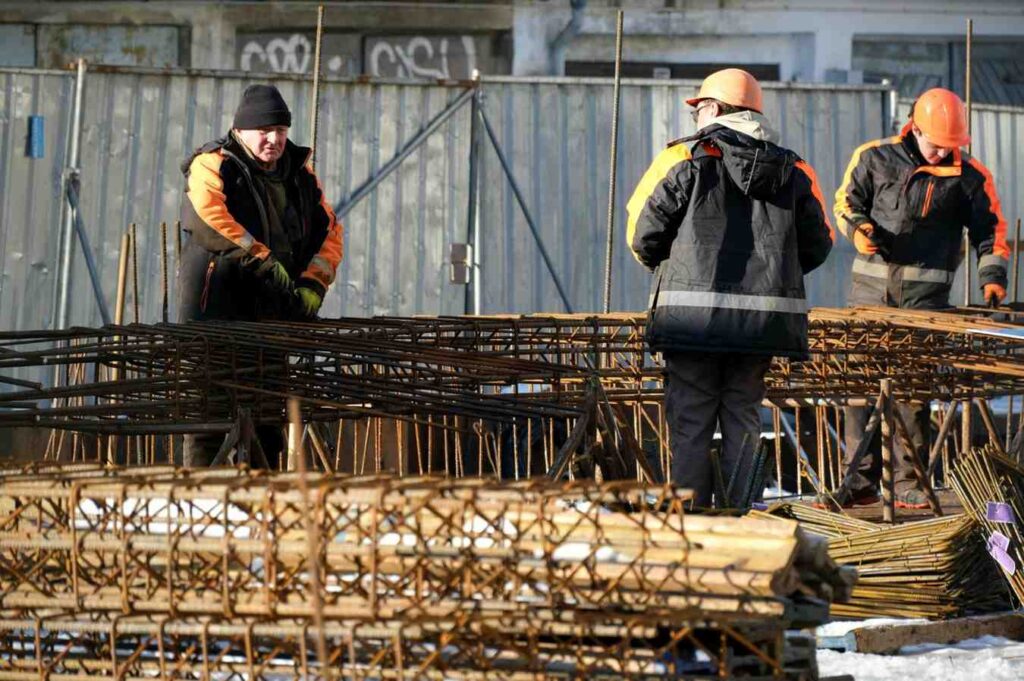 Three construction workers wearing orange safety helmets and jackets handle metal reinforcement bars at a construction site. The background shows a metal fence and a partially visible building.