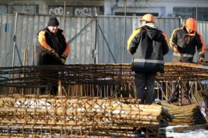 Three construction workers wearing orange safety helmets and jackets handle metal reinforcement bars at a construction site. The background shows a metal fence and a partially visible building.