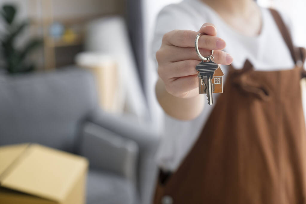 A person wearing a white shirt and brown overalls holds out a key with a small house-shaped keychain, suggesting moving into a new home. A couch and cardboard box are visible in the blurred background.