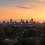 A city skyline with tall buildings is silhouetted against a vibrant orange and yellow sunset sky, with trees and houses in the foreground.