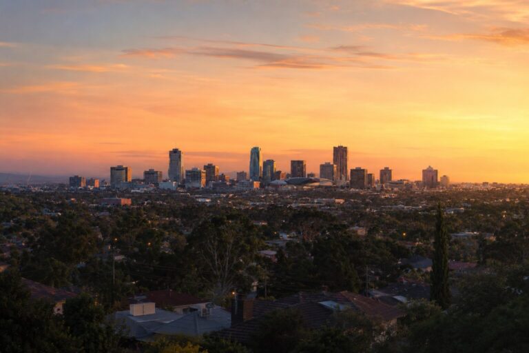 A city skyline with tall buildings is silhouetted against a vibrant orange and yellow sunset sky, with trees and houses in the foreground.