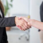 Two people wearing business attire shake hands in an office setting, suggesting a professional agreement or partnership. Only their arms and hands are visible. A person stands blurred in the background.