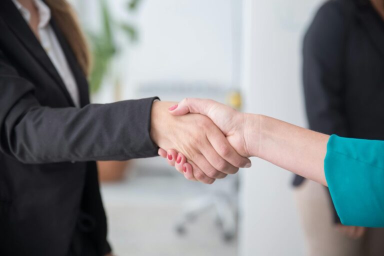 Two people wearing business attire shake hands in an office setting, suggesting a professional agreement or partnership. Only their arms and hands are visible. A person stands blurred in the background.