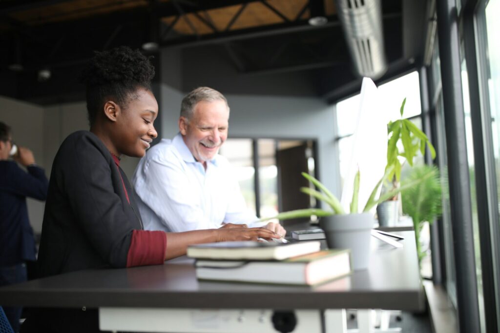 Two people sit at a desk by a large window, smiling and working together on a computer. Books and a potted plant are on the desk, and sunlight streams into the modern office space.