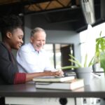 Two people sit at a desk by a large window, smiling and working together on a computer. Books and a potted plant are on the desk, and sunlight streams into the modern office space.