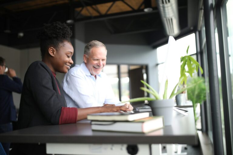 Two people sit at a desk by a large window, smiling and working together on a computer. Books and a potted plant are on the desk, and sunlight streams into the modern office space.