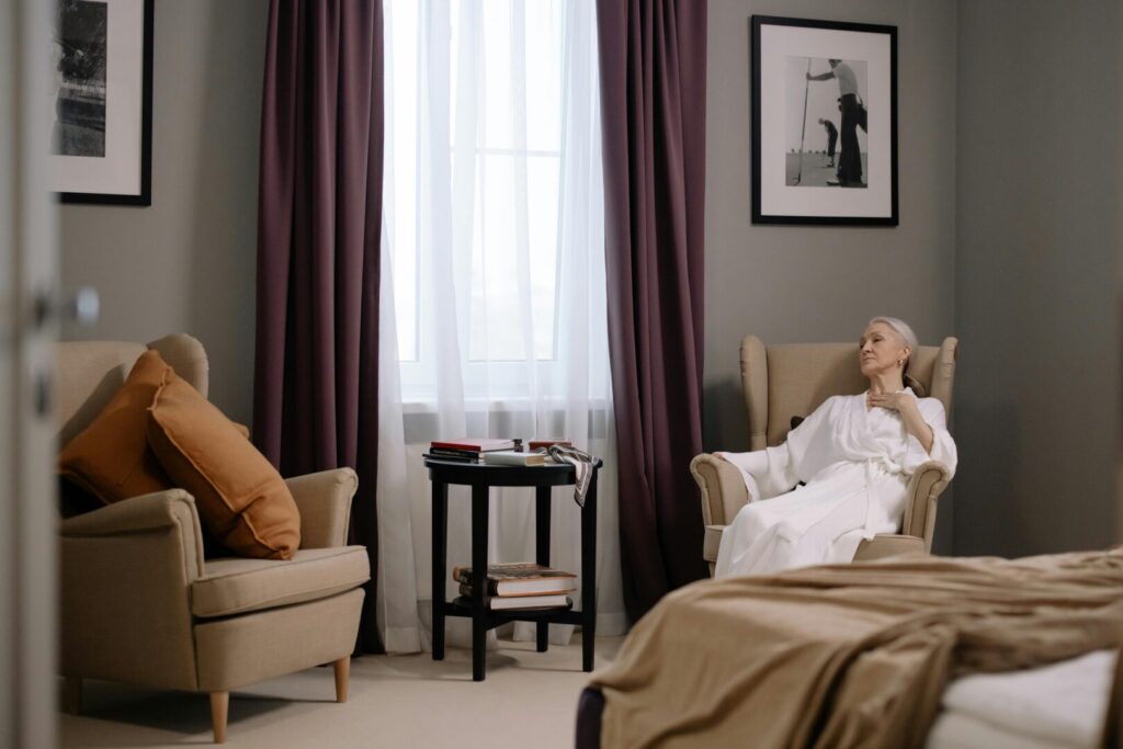 An elderly woman in a white robe sits in an armchair beside a window with purple curtains in a cozy, softly lit bedroom. A side table with books and two framed photos are visible in the background.