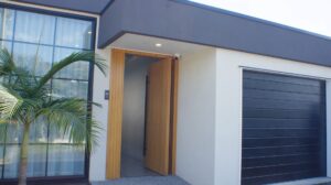 Modern house exterior with large glass window, black-framed garage door, light-colored walls, a wooden front door slightly open, and a small palm tree near the entrance.
