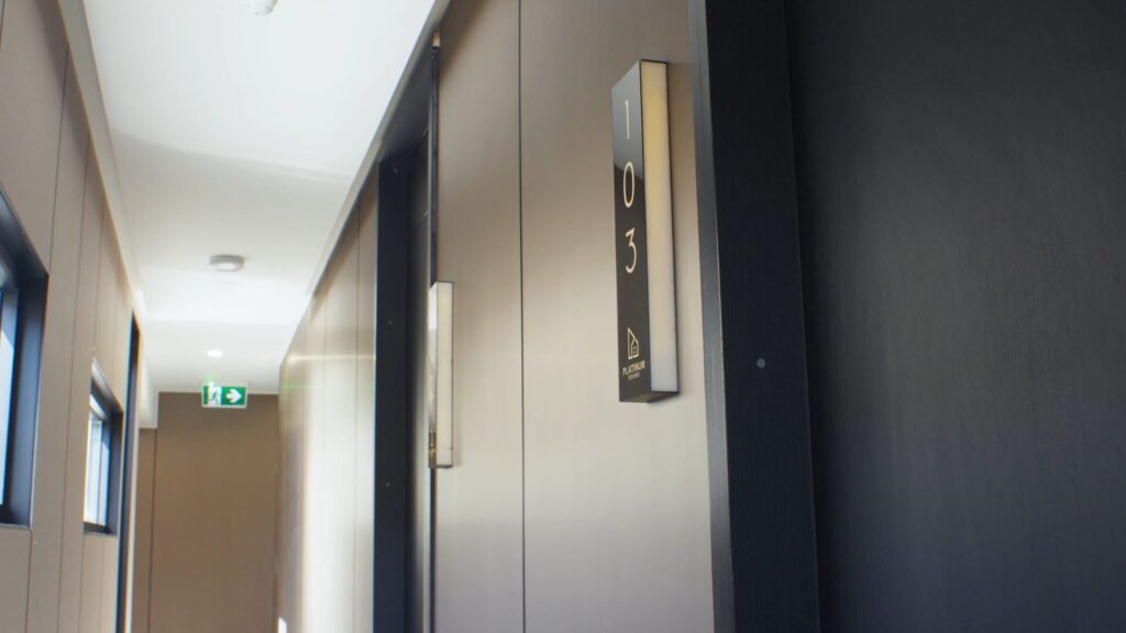 A hallway with beige walls and doors, featuring a door sign labeled 103 and an emergency exit sign visible at the end of the corridor. Natural light comes through windows on the left.
