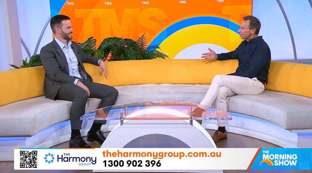 Two men sit on a yellow couch in a brightly colored TV studio, engaged in conversation. A coffee table is in front of them. The screen displays The Harmony Group website, phone number, and The Morning Show logo.