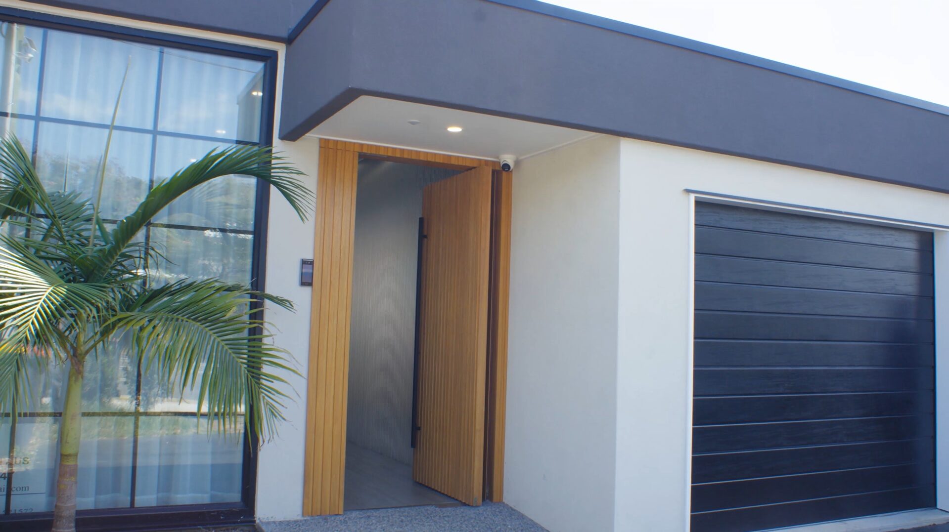 Modern house exterior with a large window, black garage door, open wooden front door, small security camera, and a palm tree by the entrance.