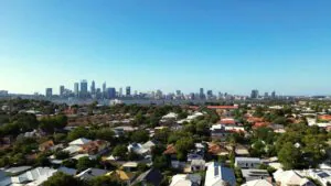 Aerial view of a suburban neighborhood with tree-lined streets and houses, with a city skyline and tall buildings in the background under a clear blue sky.