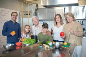 A smiling family of six, including children, adults, and a senior, stands together in a modern kitchen preparing fresh vegetables with bowls and cutting boards on the counter.