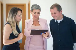 The specialist co-living property manager in the center, wearing a light pink suit, is holding the tablet and explaining something to the other woman and man, who are listening attentively.