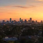 A city skyline with tall buildings is silhouetted against a vibrant orange and yellow sunset sky, with trees and houses in the foreground.