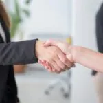 Two people wearing business attire shake hands in an office setting, suggesting a professional agreement or partnership. Only their arms and hands are visible. A person stands blurred in the background.