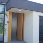 Modern house exterior with large glass window, black-framed garage door, light-colored walls, a wooden front door slightly open, and a small palm tree near the entrance.